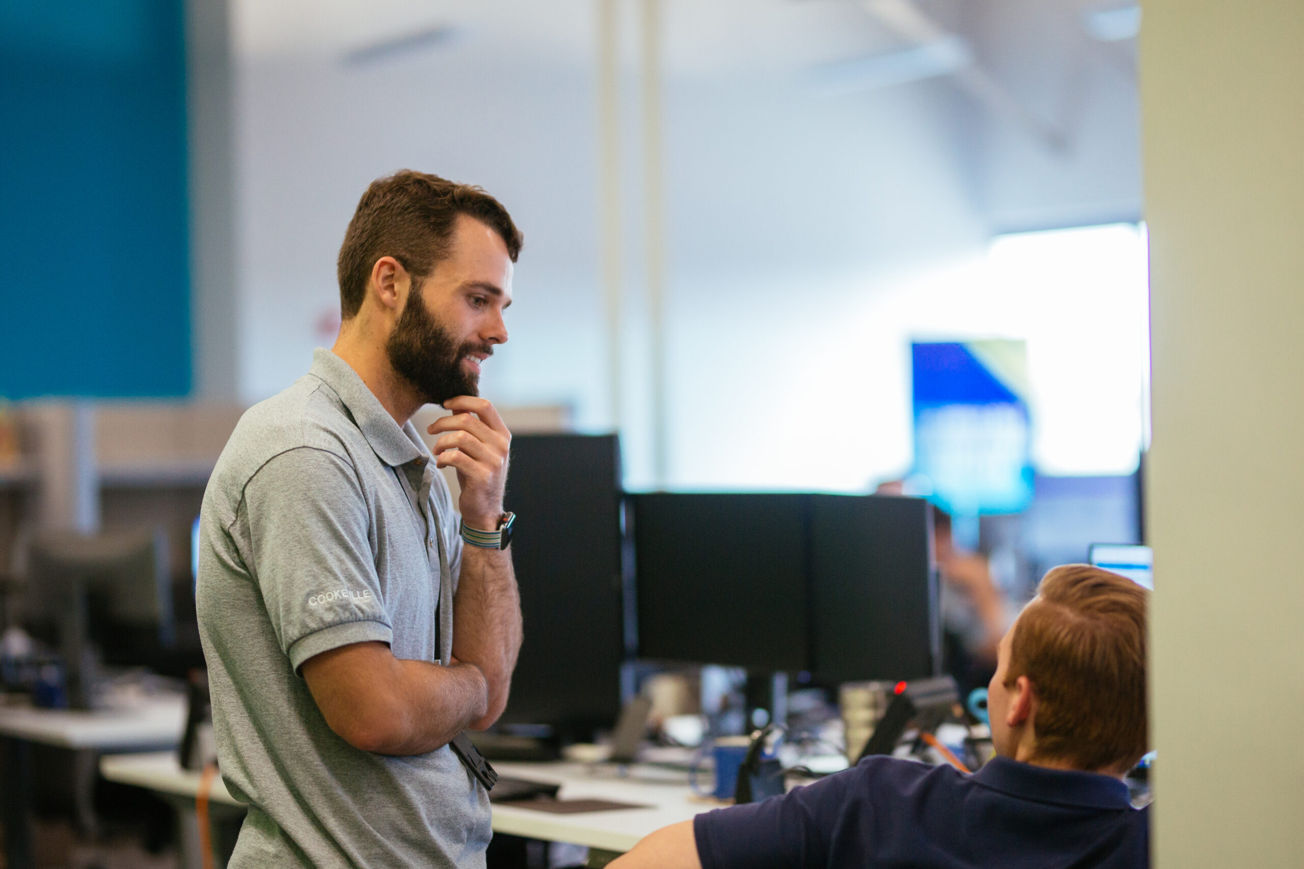 Man in gray polo talking with coworker seated at computer